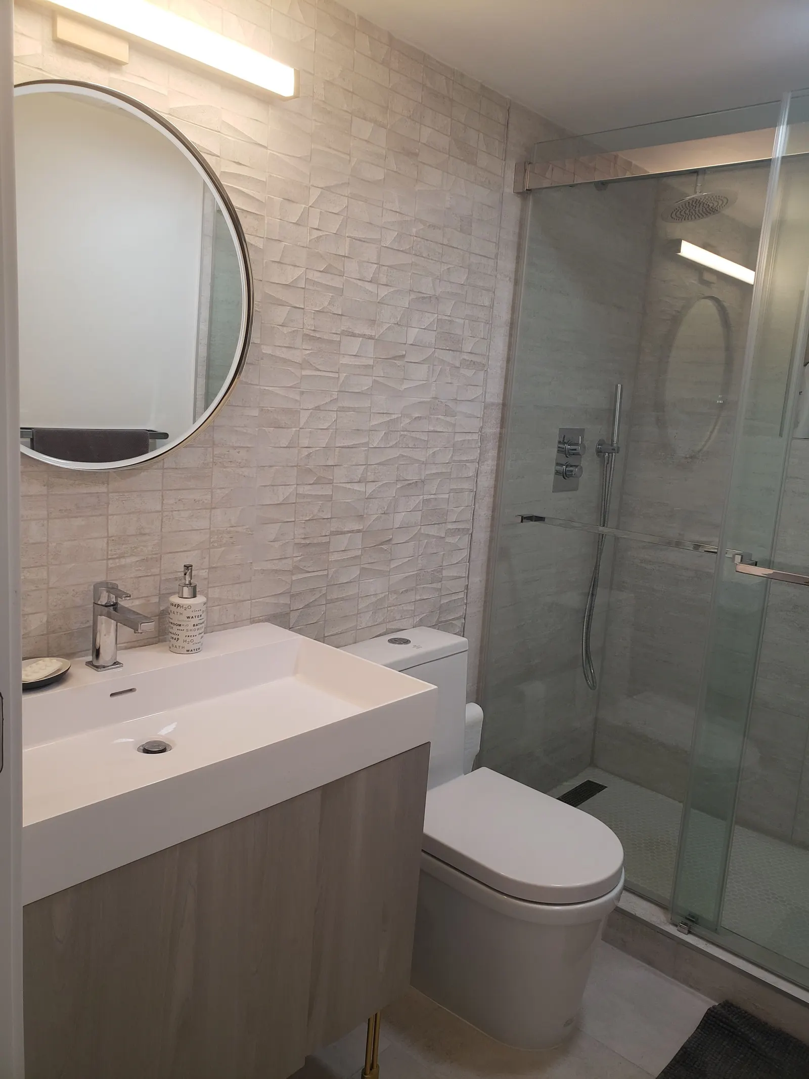 Vanidad de baño con espejo redondo, pared texturizada y ducha de vidrio